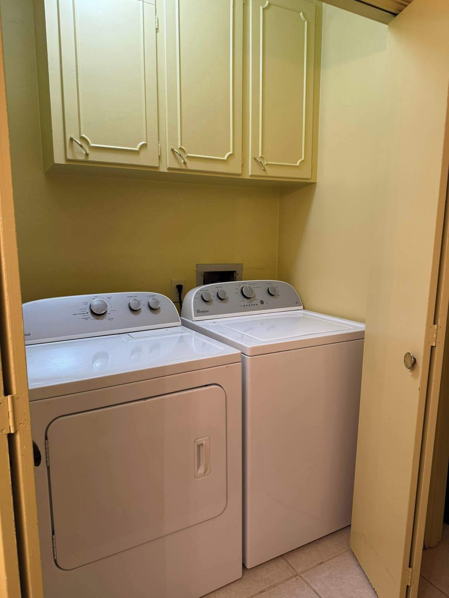 A laundry room with two white Whirlpool washers and dryers, beige cabinets above, and a door on the right.