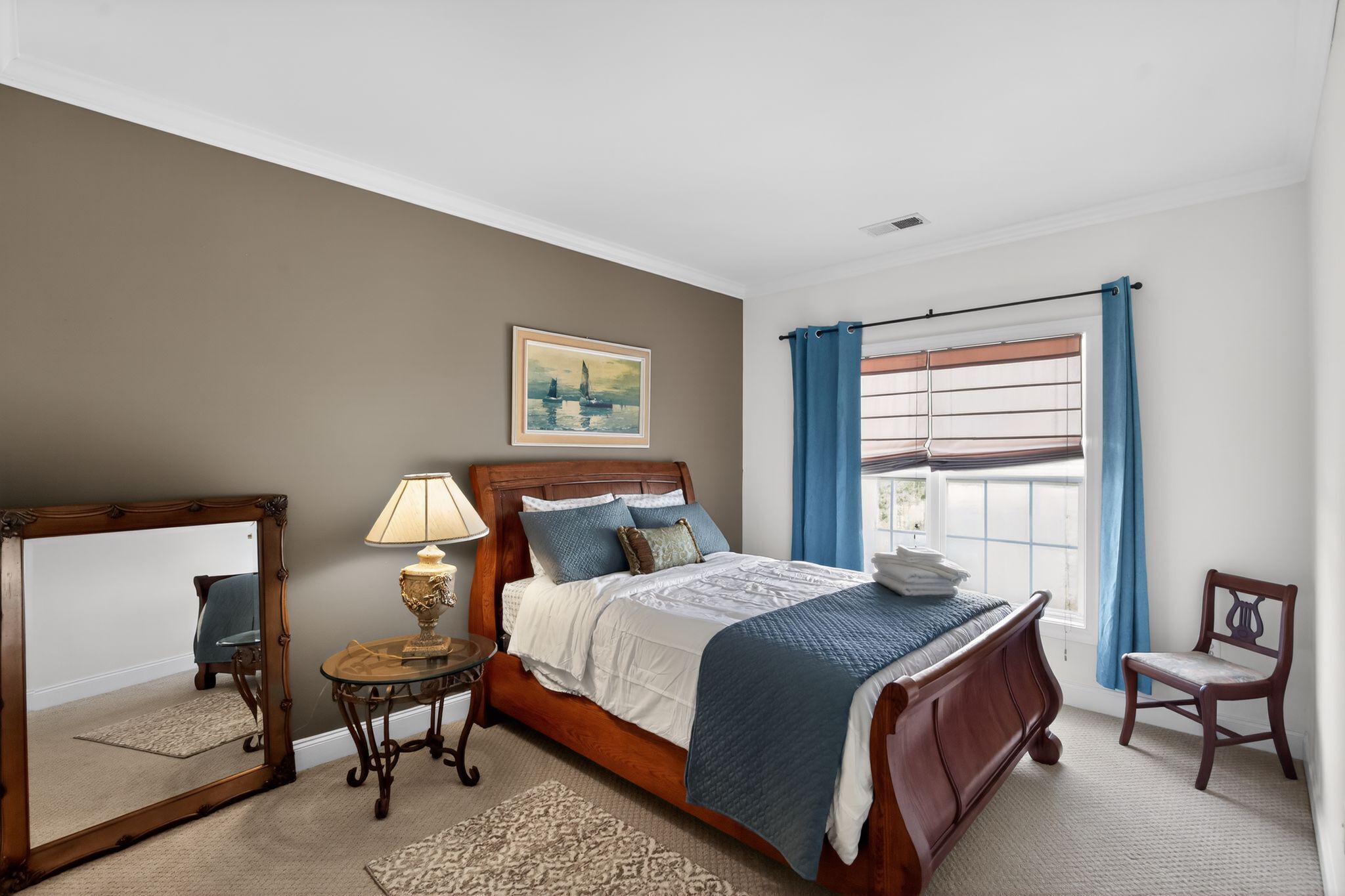 A bedroom with a bed, a mirror, a lamp, a chair, a window with curtains, and a painting on the wall.