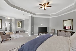 A bedroom with a bed, a television, a dresser, a chair, a ceiling fan, and a door leading to another room.