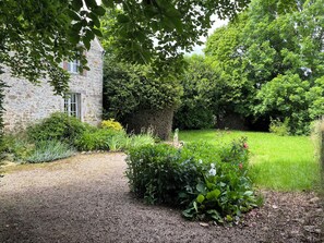 A stone building with a gravel path leading to a well-maintained garden with various plants and a hedge.