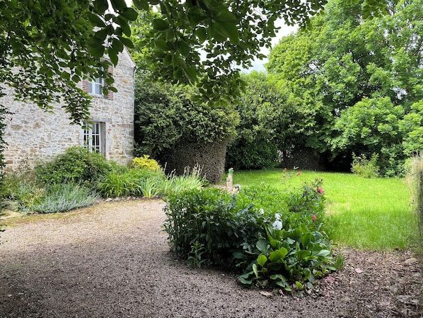 A stone building with a gravel path leading to a well-maintained garden with various plants and a hedge.