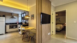Une cuisine moderne avec un coin repas, un téléviseur fixé sur un mur en bois, et une chambre à coucher visible à travers une porte ouverte.