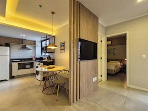 A modern kitchen with a dining area, a TV mounted on a wooden partition, and a bedroom visible through an open door. - Apartment 13 - Jundiaí (Jundiaí)