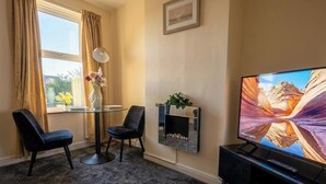 Habitación de hotel con televisión, una pequeña mesa con sillas, un florero con flores y una planta en un estante.