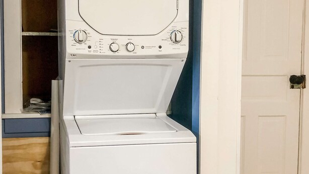 A front-loading washing machine in a laundry room with a wooden floor and a white door. - The Three Seasons | Centrally located house in Lak (Lake Leelanau)