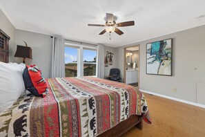 Um quarto com cama, ventilador de teto, janela, cadeira e um quadro na parede.