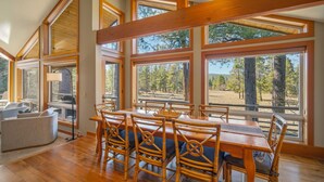 Un comedor con mesa y sillas de madera, ventanas amplias con vistas al bosque y un techo alto con vigas a la vista.