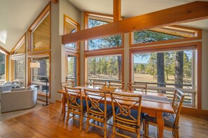 A dining area with a wooden table and chairs, large windows with a forest view, and a high ceiling with exposed beams. - Spacious 4bd/4bath Riverfront Retreat in Sunriver (Sunriver)