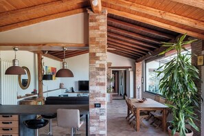A rustic interior with exposed wooden beams, a brick column, a dining area with a large plant, and a living room with a sofa and TV. - "Sunrise over the sea and relaxation on Etna: Green oasis near the pearls of Sicily. (Sicilia)