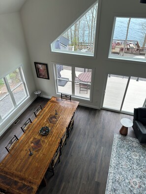 A spacious room with a large wooden dining table, multiple chairs, and large windows offering views of a deck and outdoor area.