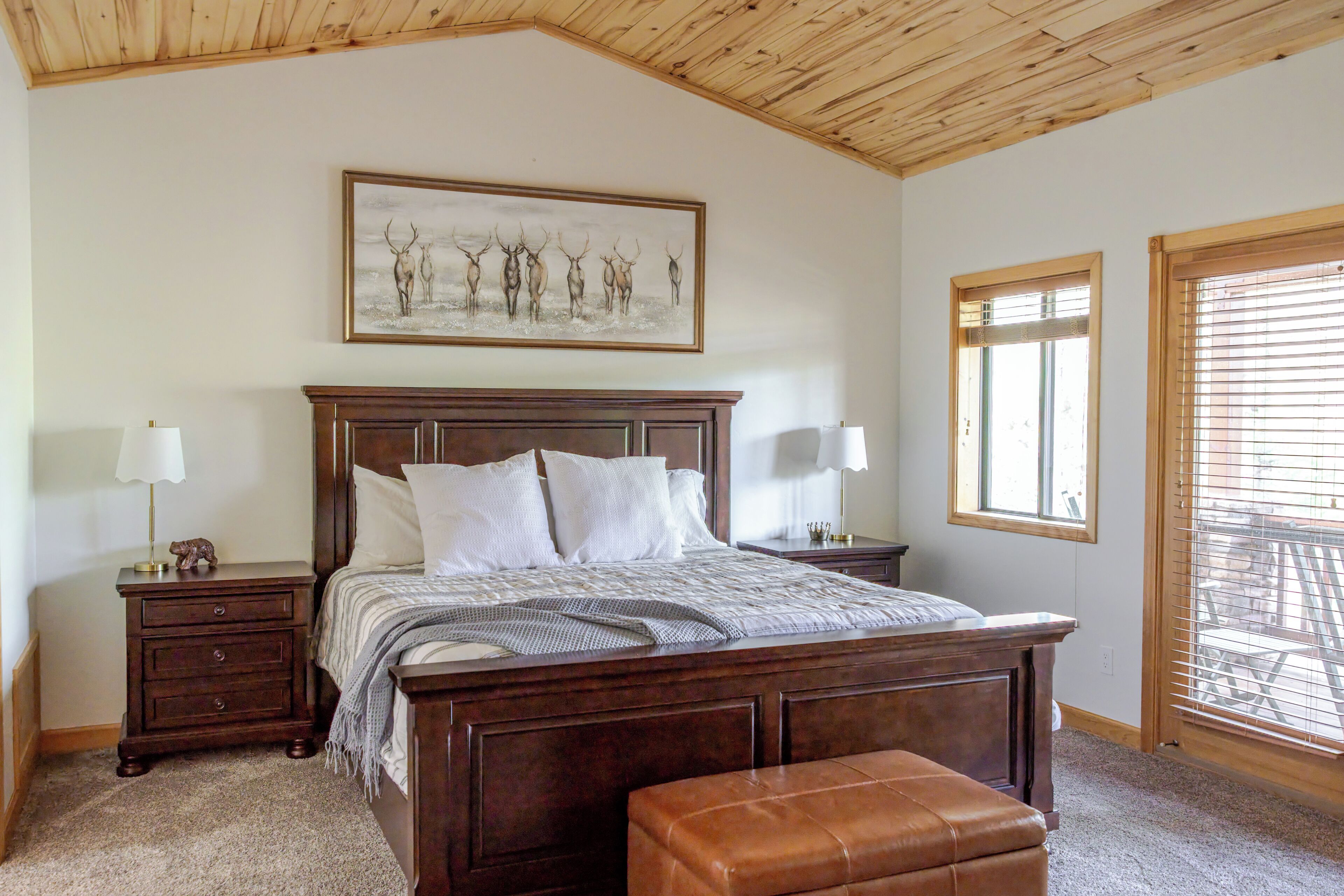 Un dormitorio con techo de madera, una cama grande con cabecera, dos mesitas de noche con lámparas, un banco y una ventana con persianas.