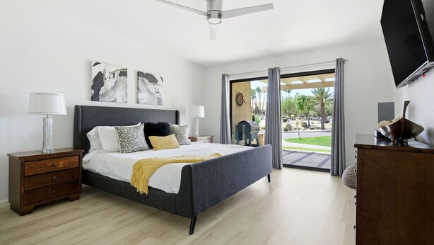 A modern bedroom with a bed, nightstands, a ceiling fan, a TV, and a view of the outdoors. - 10th Tee Retreat - Mountain Views + Resort Living (Palm Desert)