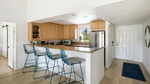 A modern kitchen with wooden cabinets, a stainless steel refrigerator, and a granite countertop. There are three blue bar stools and a window providing a view of the outdoors. - 10th Tee Retreat - Mountain Views + Resort Living (Palm Desert)