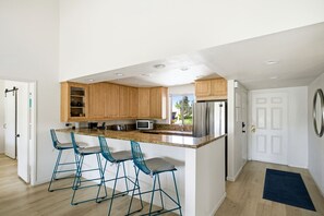 A modern kitchen with wooden cabinets, a stainless steel refrigerator, and a granite countertop. There are three blue bar stools and a window providing a view of the outdoors. - 10th Tee Retreat - Mountain Views + Resort Living (Palm Desert)