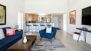 A modern living room with a sofa, coffee table, and a blue armchair. A kitchen with bar stools is visible in the background. - 10th Tee Retreat - Mountain Views + Resort Living (Palm Desert)