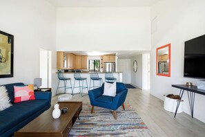 A modern living room with a sofa, coffee table, and a blue armchair. A kitchen with bar stools is visible in the background. - 10th Tee Retreat - Mountain Views + Resort Living (Palm Desert)