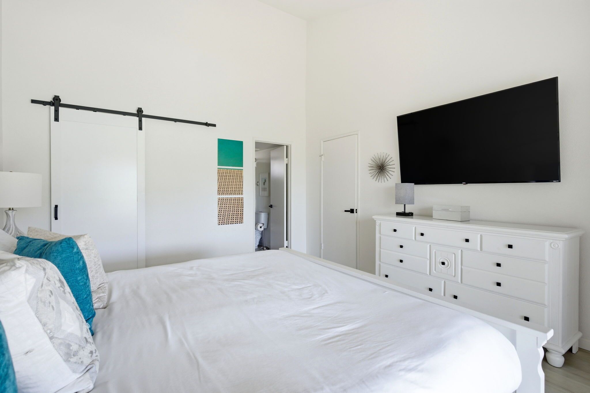 A bedroom with a bed, a dresser, a TV, a door, and a wall-mounted shelf.