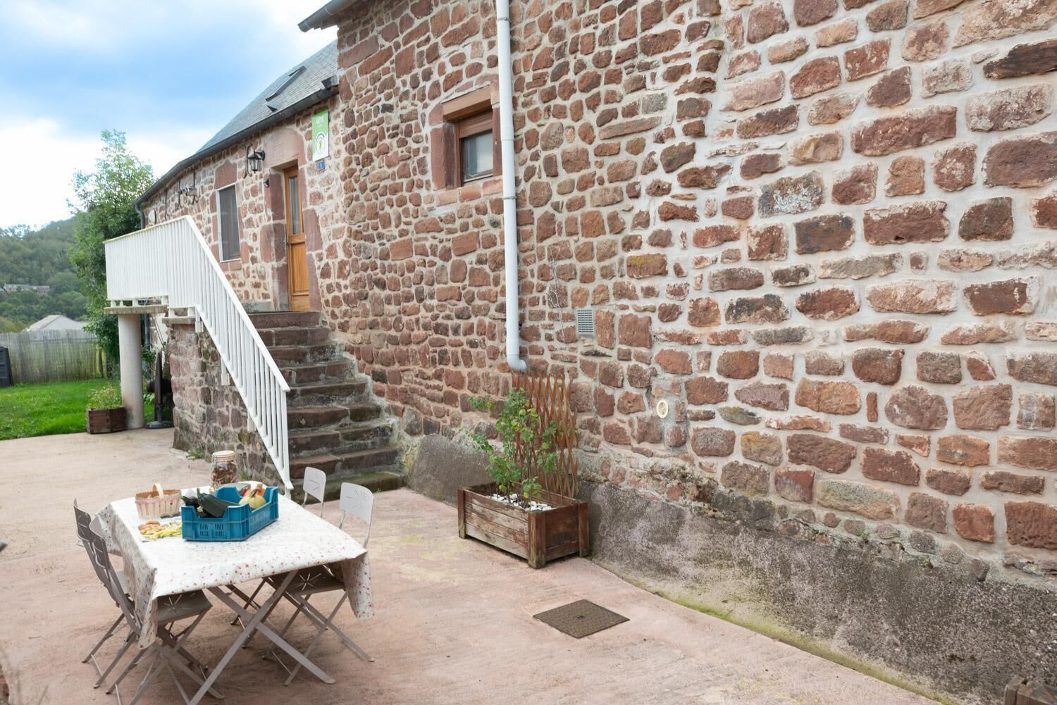 A stone building with a staircase leading to a door, a table with chairs outside, and a small plant in a wooden box.