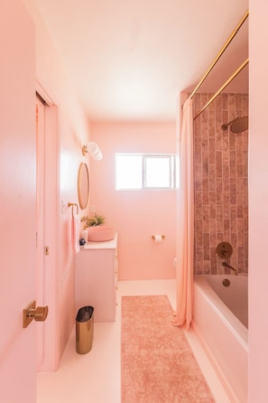 Une salle de bain aux couleurs roses, avec une baignoire, un lavabo avec un miroir et une fenêtre.