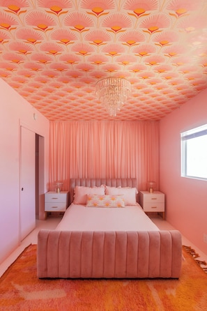 A pink-themed bedroom with a patterned ceiling, a chandelier, a bed, nightstands, and a window.