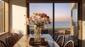 Ein Esstisch mit Stühlen, eine Blumenvase und Blick auf das Meer durch große Fenster.
