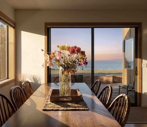 A dining table with chairs, a vase of flowers, and a view of the ocean through large windows. - Stunning Oceanfront Home with 3 Ensuites + Hot Tub (Sea Ranch)