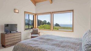 Ein Schlafzimmer mit einem großen Bett, einem Fernseher auf einer Holzkommode und einem Sessel. Drei große Fenster bieten einen Blick ins Freie.