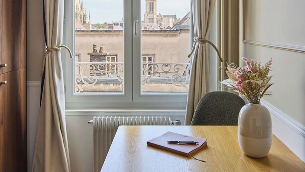 A room with a large window, a table with a notebook and pen, a vase with flowers, and a radiator. - Le Carrousel - Haussmann hyper-centre Troyes - Quiet and rooftop views (Troyes)