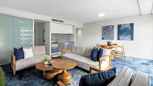 A modern living room with a sofa, coffee table, and chairs, a kitchen area, and abstract wall art. - Waterfront at Harbour Light—Generous Space & Views (Cairns)