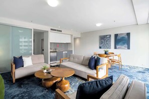 A modern living room with a sofa, coffee table, and chairs, a kitchen area, and abstract wall art. - Waterfront at Harbour Light—Generous Space & Views (Cairns)