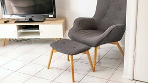 Un salon moderne avec un fauteuil gris, un pouf assorti, un téléviseur à écran plat posé sur un meuble en bois et un sol carrelé de couleur blanche.