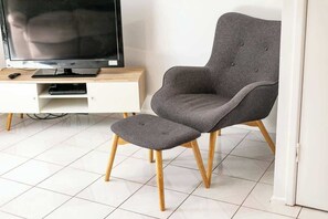 Ein modernes Wohnzimmer mit einem grauen Sessel, passendem Hocker, einem Flachbildfernseher auf einem Holzboden und einem weißen Fliesenboden.