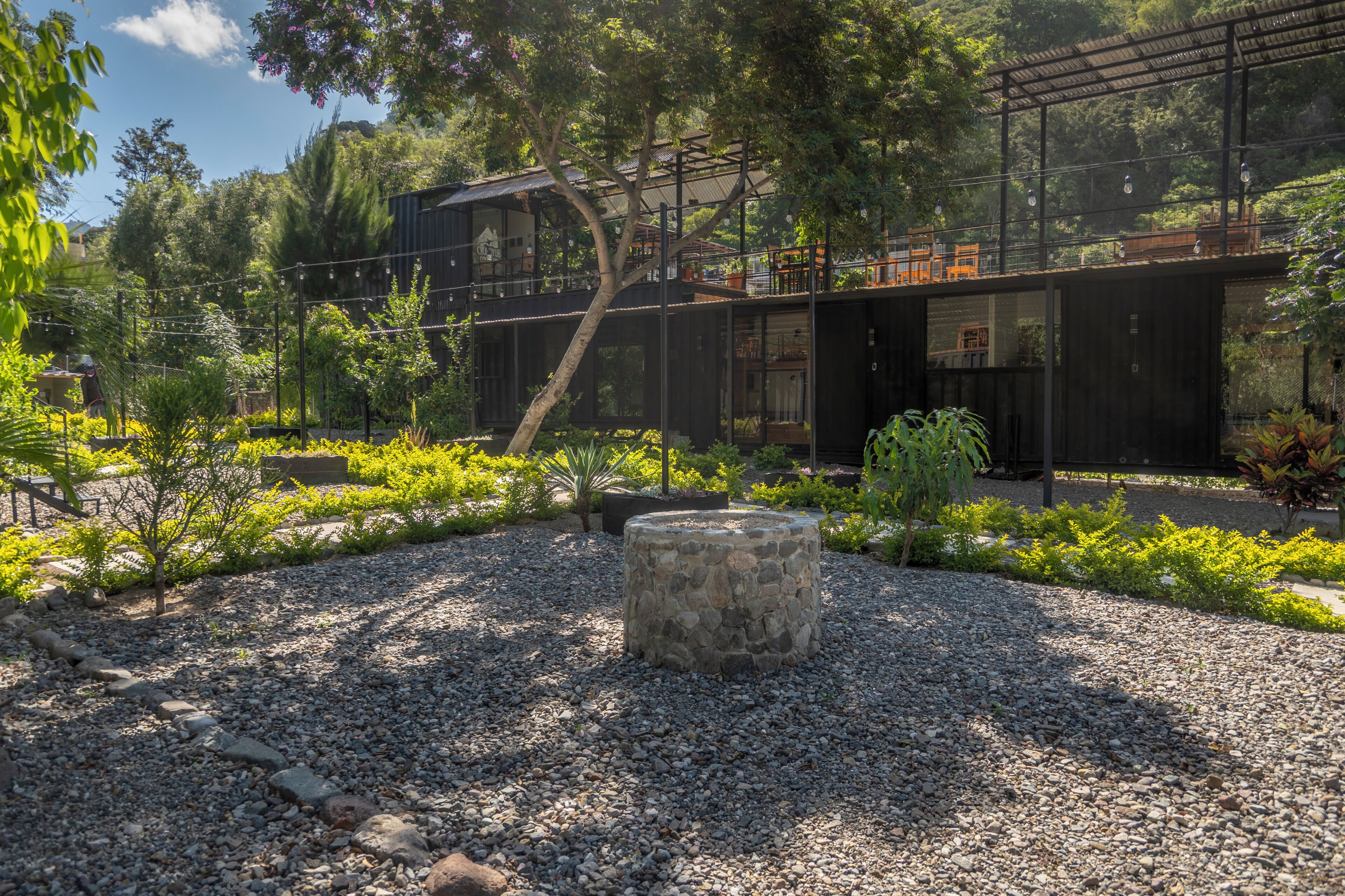 Un espacio exterior moderno con una jardinera de piedra, suelo de grava y un edificio con techo metálico.