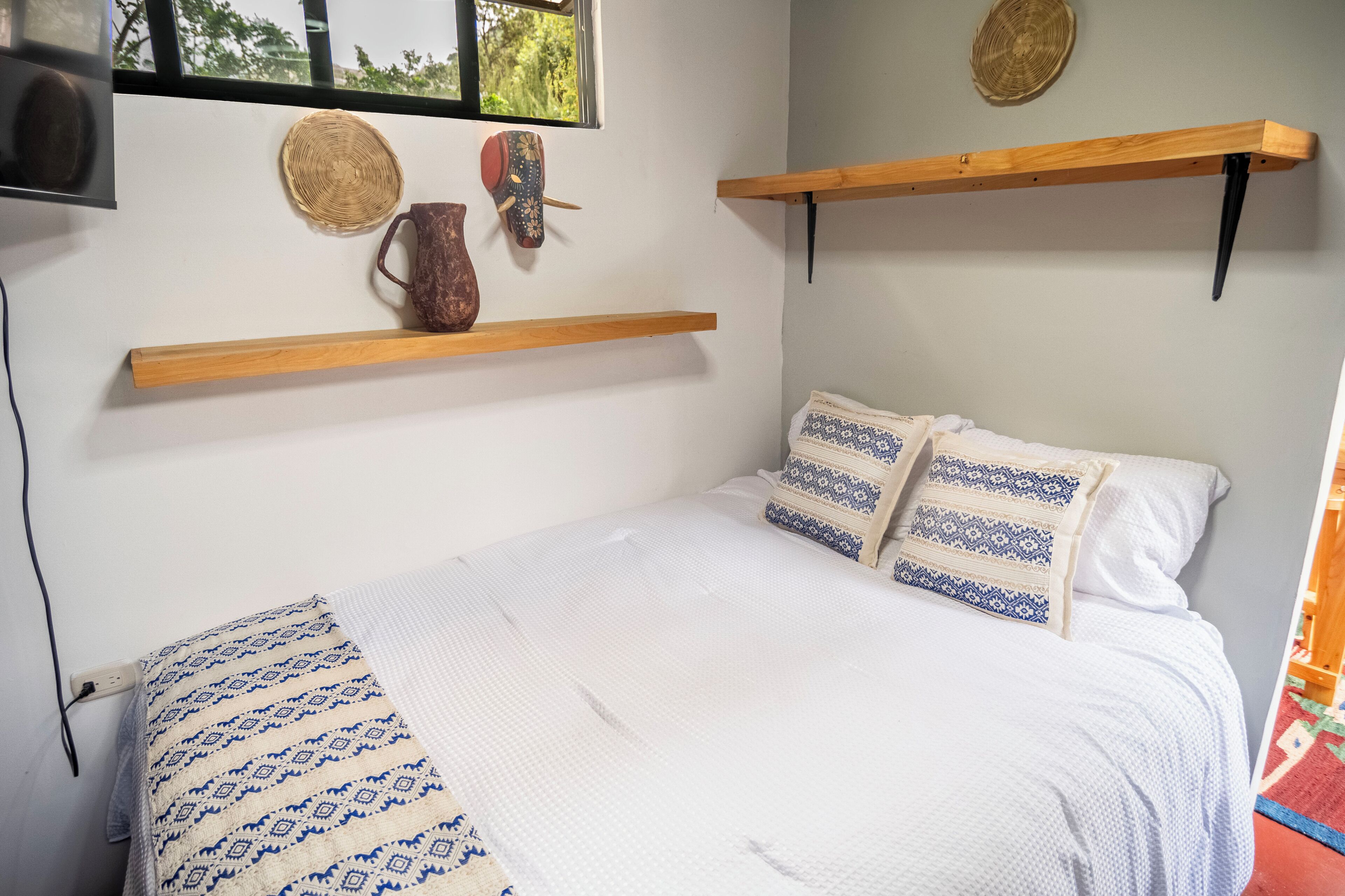 Un dormitorio con cama, almohadas, una mesita de noche y estantes en la pared.