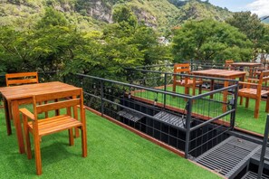 Un área de estar al aire libre con mesas y sillas de madera, rodeada por una barandilla metálica, con vista a césped y árboles verdes.