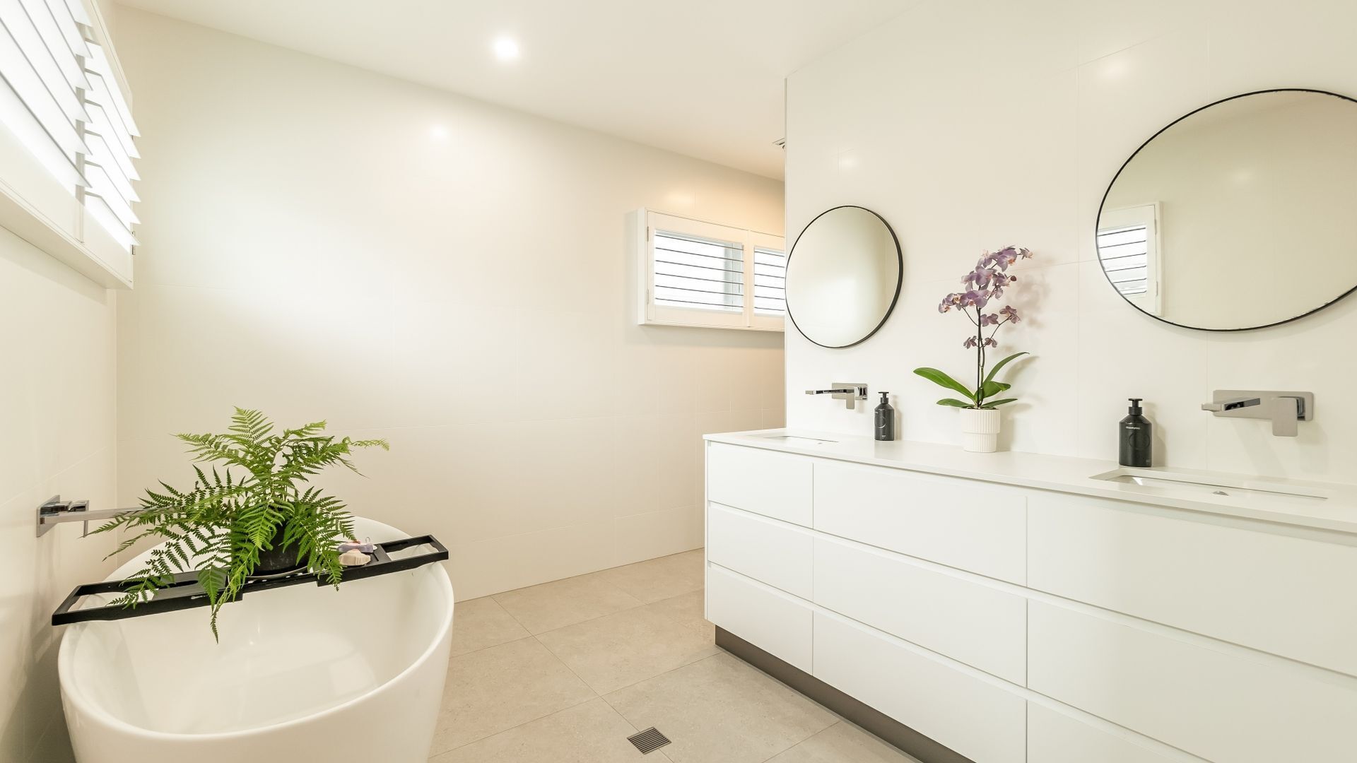 A modern bathroom with a freestanding bathtub, two wall-mounted mirrors, a white vanity with a sink, and a potted orchid.
