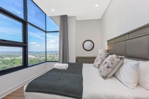 Ein Schlafzimmer mit großem Fenster, einem Bett, einem Nachttisch und Blick auf die Stadt.