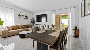 Una sala de estar moderna con un sofá, una mesa de centro, una mesa de comedor y sillas. Hay un televisor montado en la pared y una planta al lado.