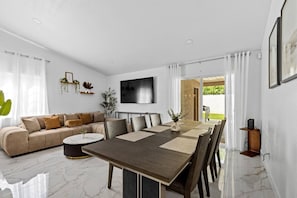 A modern living room with a sofa, coffee table, dining table, and chairs. There is a TV mounted on the wall and a plant on the side. - Spacious 7BR/5BA 8 Beds w/Hot Tub | Near & Airport (Las Vegas)