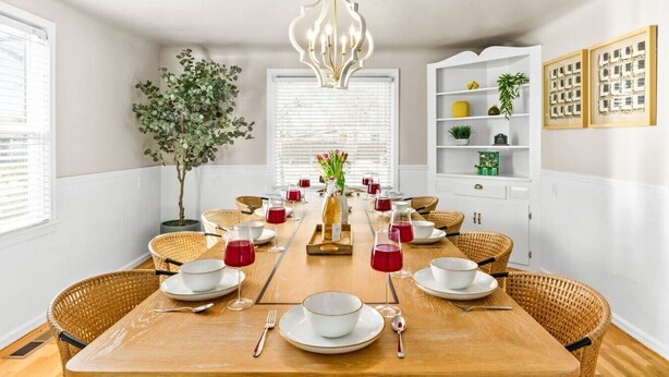 Ein Esszimmer mit einem langen Tisch, der für eine Mahlzeit gedeckt ist, umgeben von Korbstühlen, mit einem Chandelier darüber und einer Topfpflanze links.