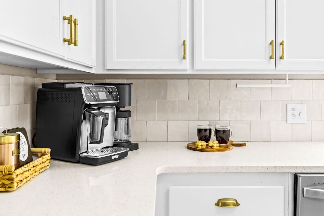 Uma cozinha com armários brancos, uma máquina de café, duas xícaras de café, uma cesta com um recipiente e um pequeno eletrodoméstico.