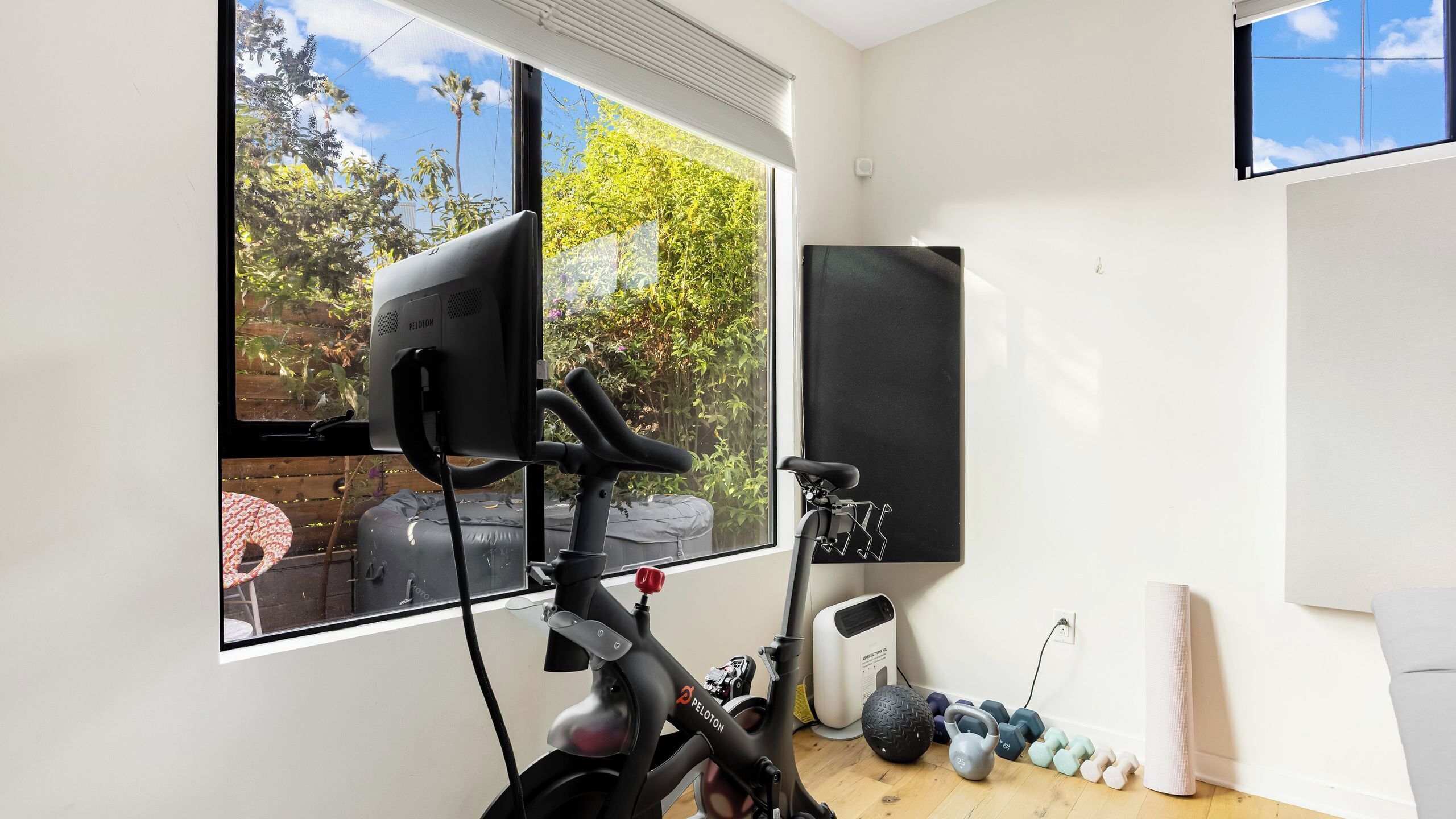Une pièce équipée d’un vélo d’exercice Peloton, d’une grande fenêtre donnant sur un espace verdoyant, d’un téléviseur fixé au mur et d’un ensemble d’haltères.