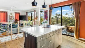 A modern kitchen with a large island, a dining area with a table and chairs, and a balcony with a view of palm trees. - Silverlake Sunset Treehouse Art-filled Escape w/ Jacuzzi + Sauna Avantstay (Los Angeles)