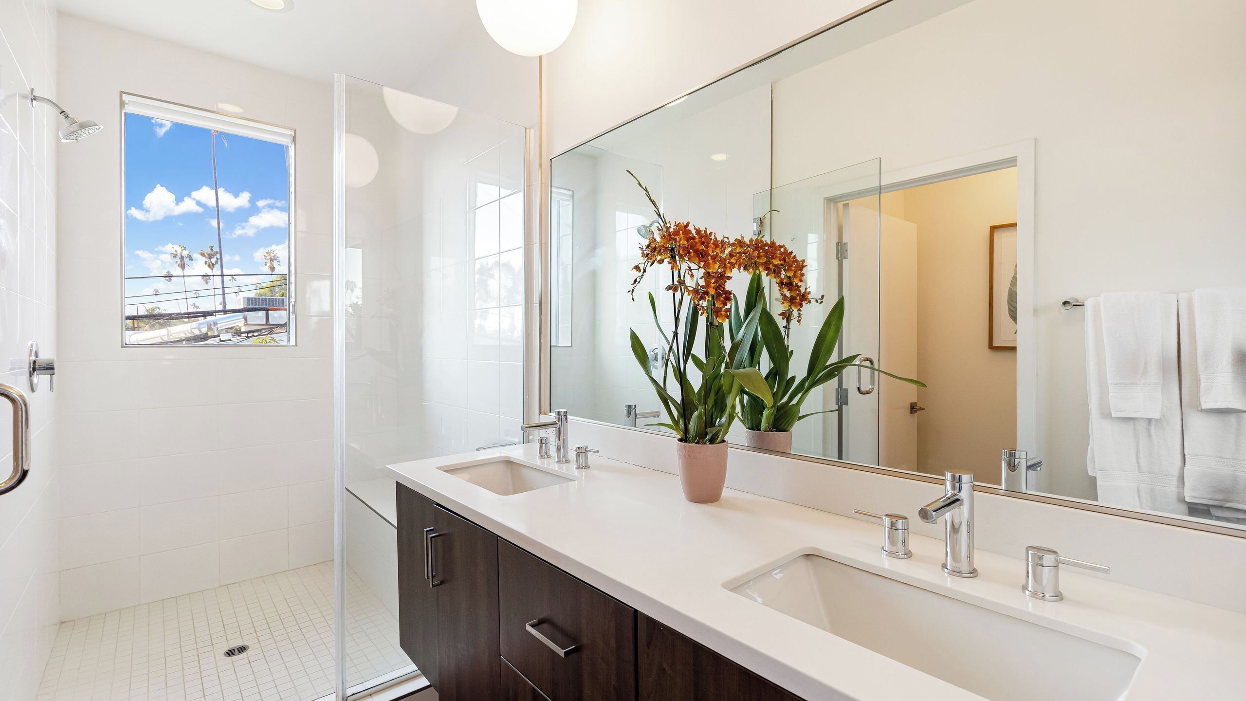 Un baño moderno con un espejo grande, dos lavamanos, una ducha y una ventana con vista a un edificio.