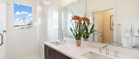 Un baño moderno con un espejo grande, dos lavamanos, una ducha y una ventana con vista a un edificio.