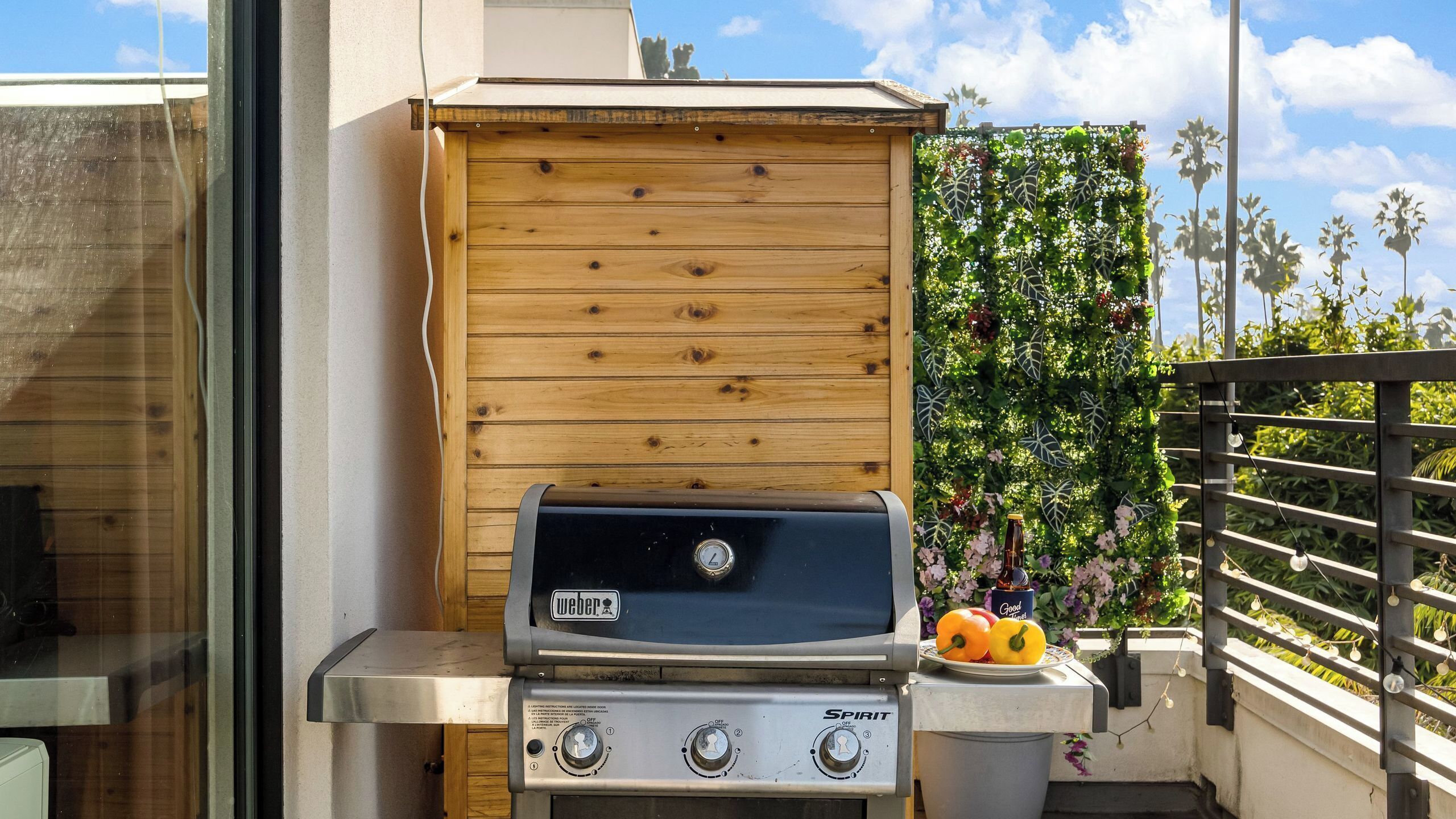 Een draagbare Weber barbecue met een houten kast, een kleine vuilnisbak en een decoratieve muur met planten en bloemen.