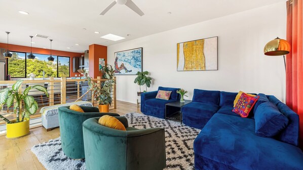 A modern living room with a blue sofa set, green armchairs, and a black coffee table. There are two large paintings on the wall, a ceiling fan, and a view of greenery through large windows.