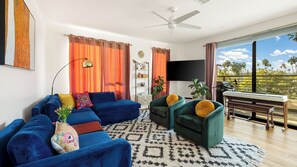 Una sala moderna con un sofá azul, un sillón verde, un piano y un amplio ventanal con vistas a palmeras.