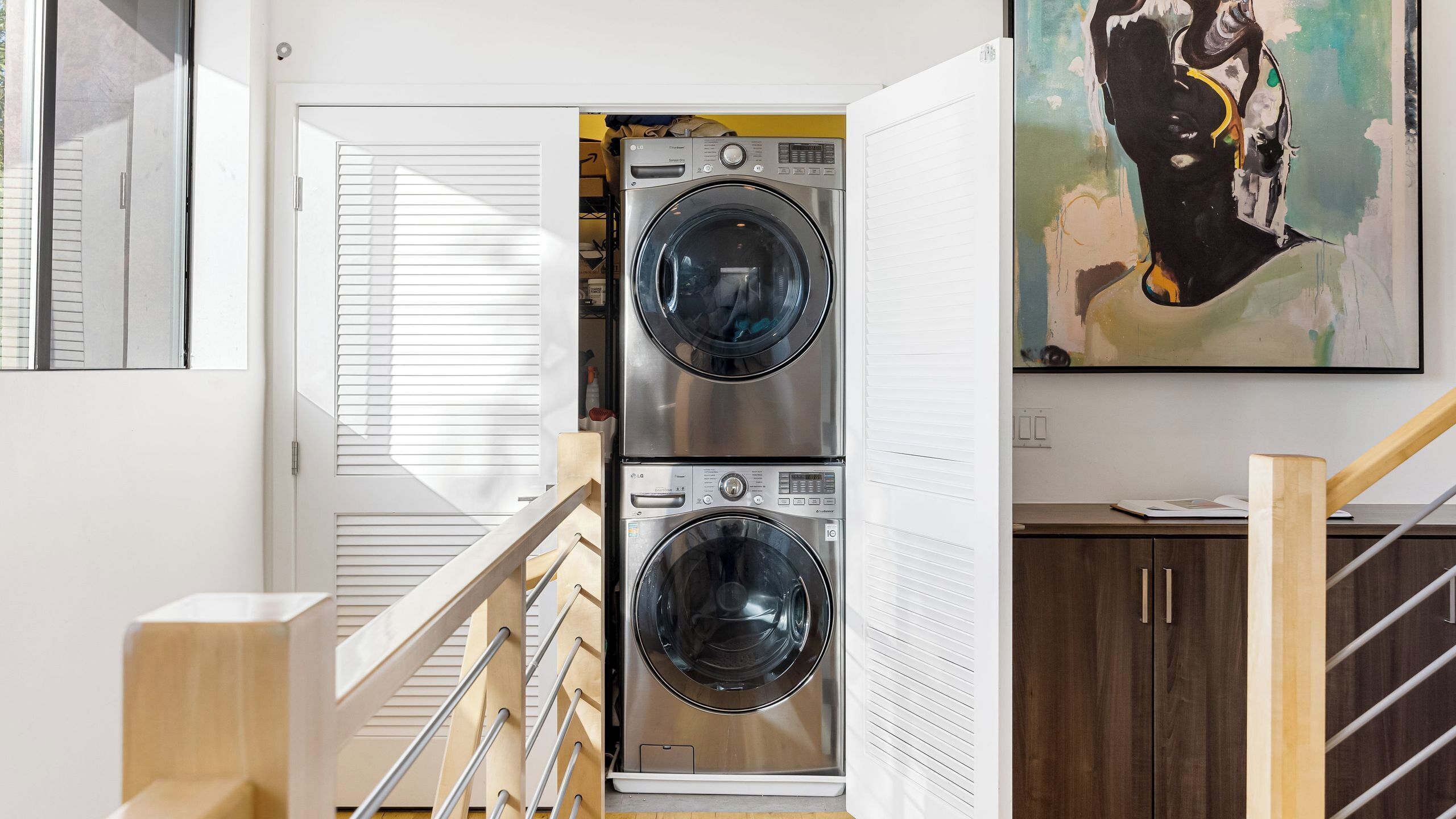 A laundry room with two stacked front-loading washing machines, a wooden staircase, and a painting on the wall.