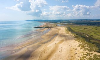 Vista aérea de una zona costera con playa arenosa, una carretera curva y campos verdes distantes.